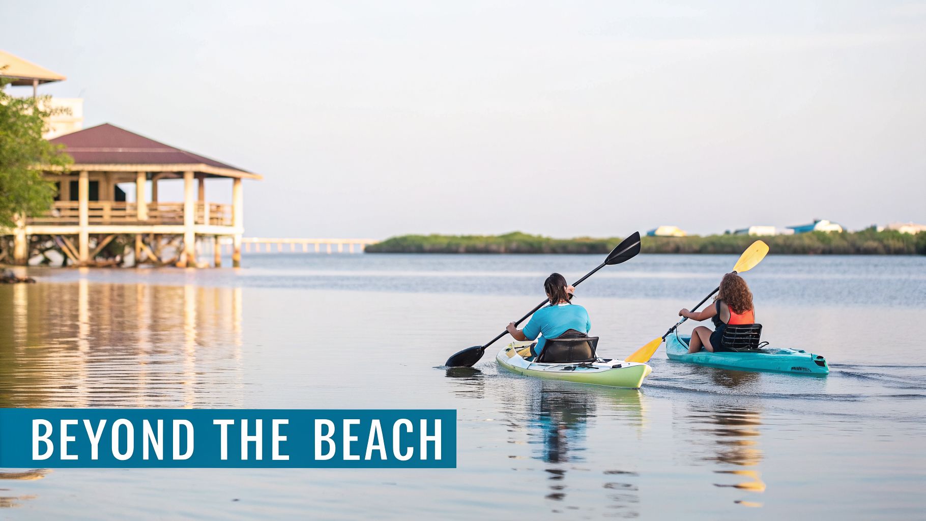 Two people kayaking on calm water with a stilt house and a distant bridge in the background.